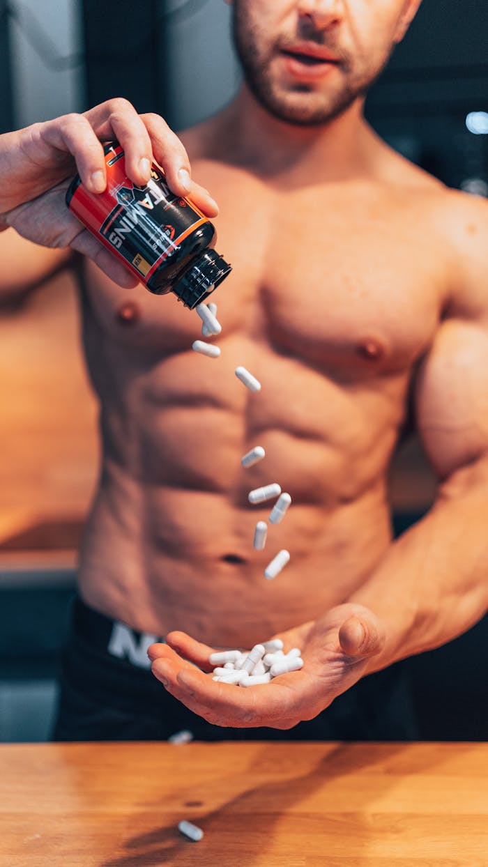 about-04 Shirtless man pouring white supplement capsules into his hand with muscles in focus.